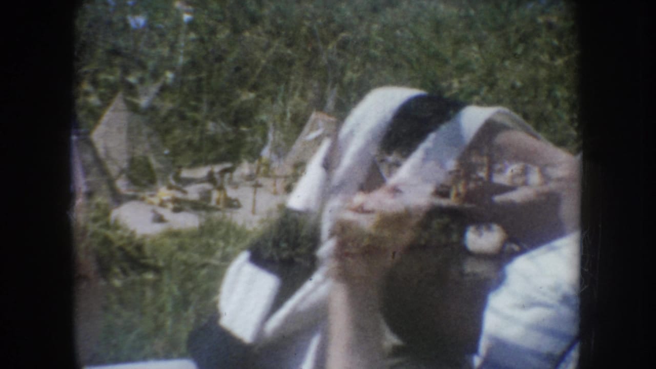 Young girl in light dress beside outdoor tipi display structures, green foliage behind, likely South Dakota, c.1955, 8mm home movie.