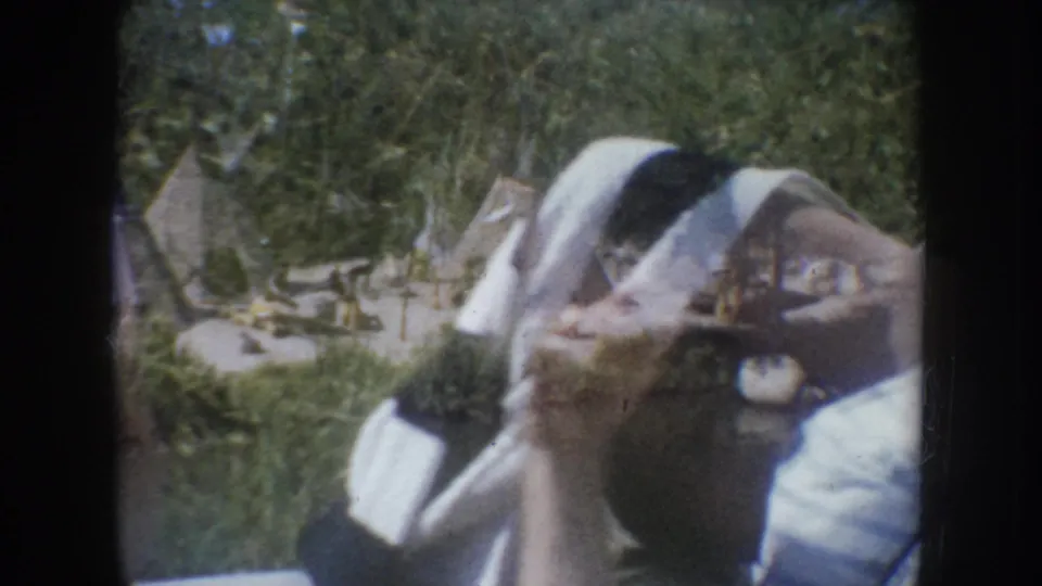 Young girl in light dress beside outdoor tipi display structures, green foliage behind, likely South Dakota, c.1955, 8mm home movie.