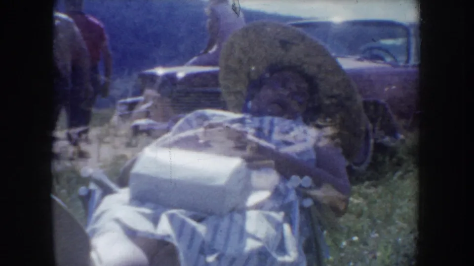Outdoor tourist attraction with circular display element in foreground, a large-finned 1950s automobile parked behind, and adult figures visible, South Dakota c.1955, 8mm home movie.