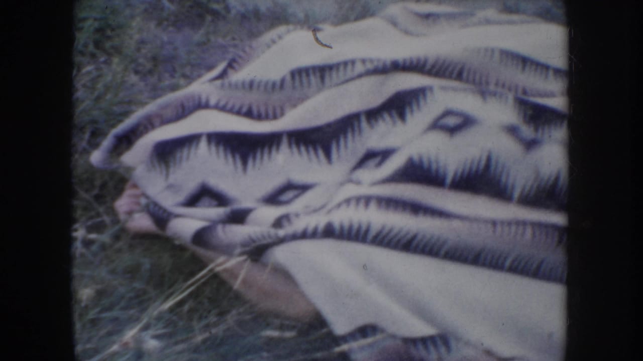 Close-up of a patterned camp-style blanket with diamond and chevron motifs on grass, human figure partially hidden beneath it, 1950s 8mm home movie.