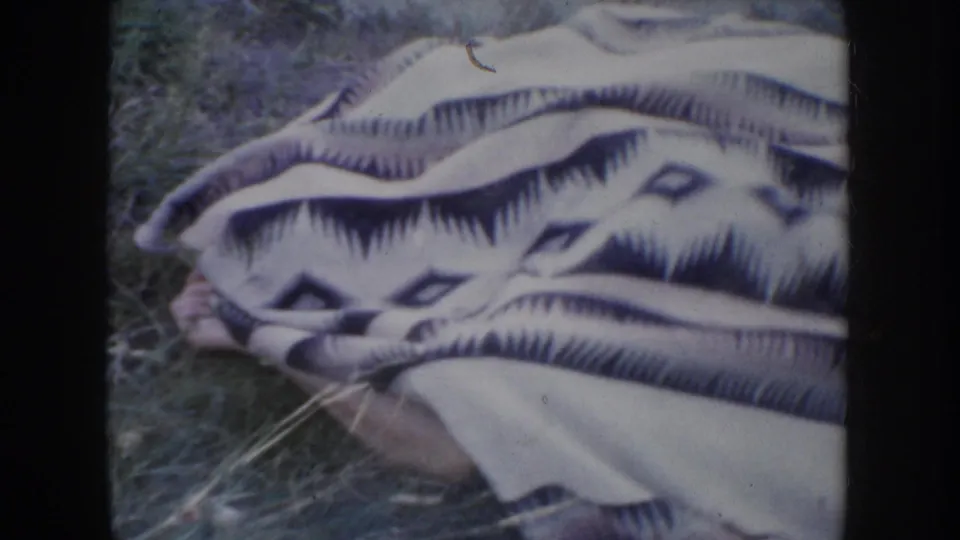 Close-up of a patterned camp-style blanket with diamond and chevron motifs on grass, human figure partially hidden beneath it, 1950s 8mm home movie.