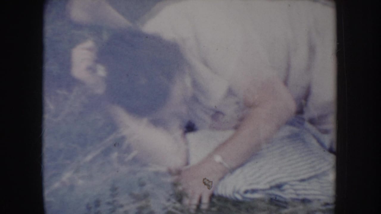 Man in light short-sleeved shirt bent forward, face angled down, bracing a hand on grass as he pushes himself upright; patterned blanket visible at right.