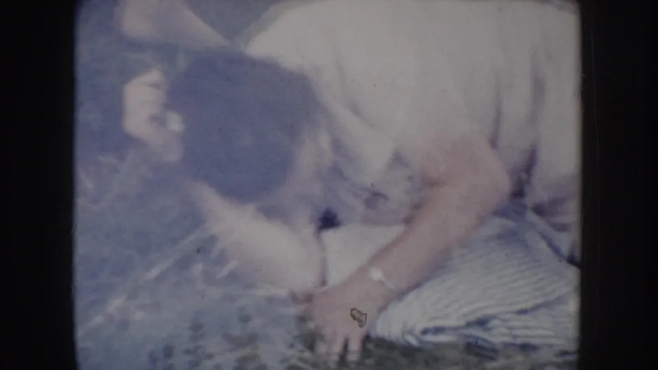 Man in light short-sleeved shirt pushing himself up from the ground on summer grass, face not clearly visible; patterned blanket at right edge, circa 1955 8mm home movie.