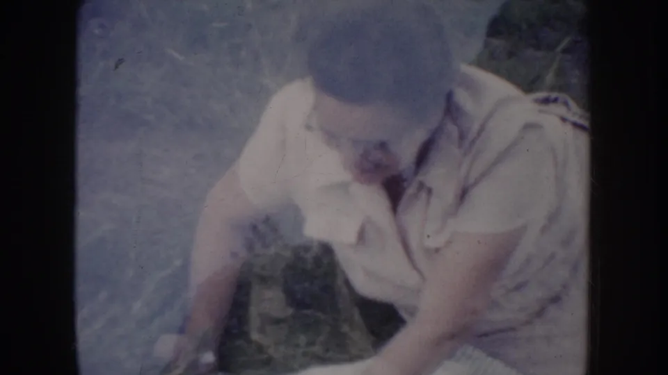 Man in light short-sleeved collared shirt seated on dry summer grass, face angled downward, gathering white fabric or blanket, 1950s 8mm home movie.