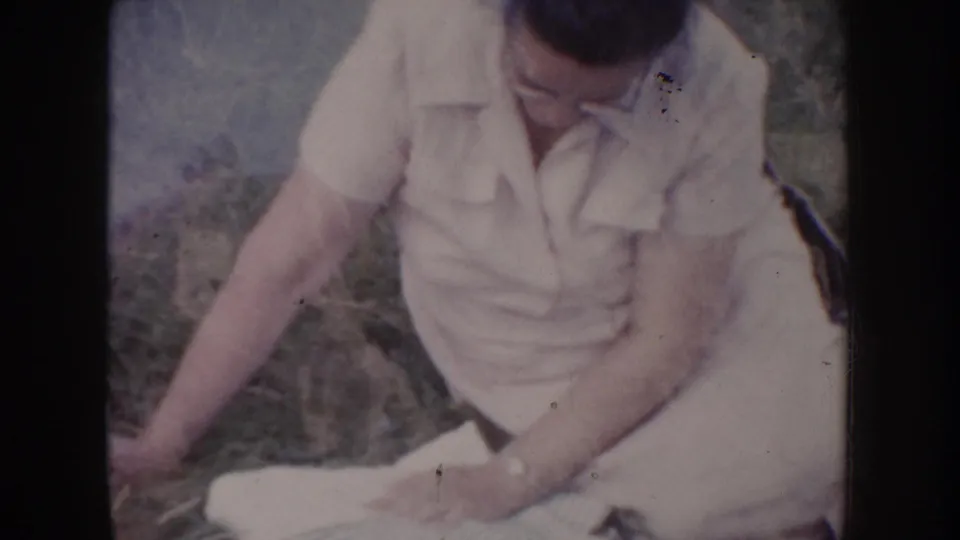 Man in white short-sleeved collared shirt sitting up on dry grass and holding white fabric, face partially visible, South Dakota circa 1955 8mm home movie.