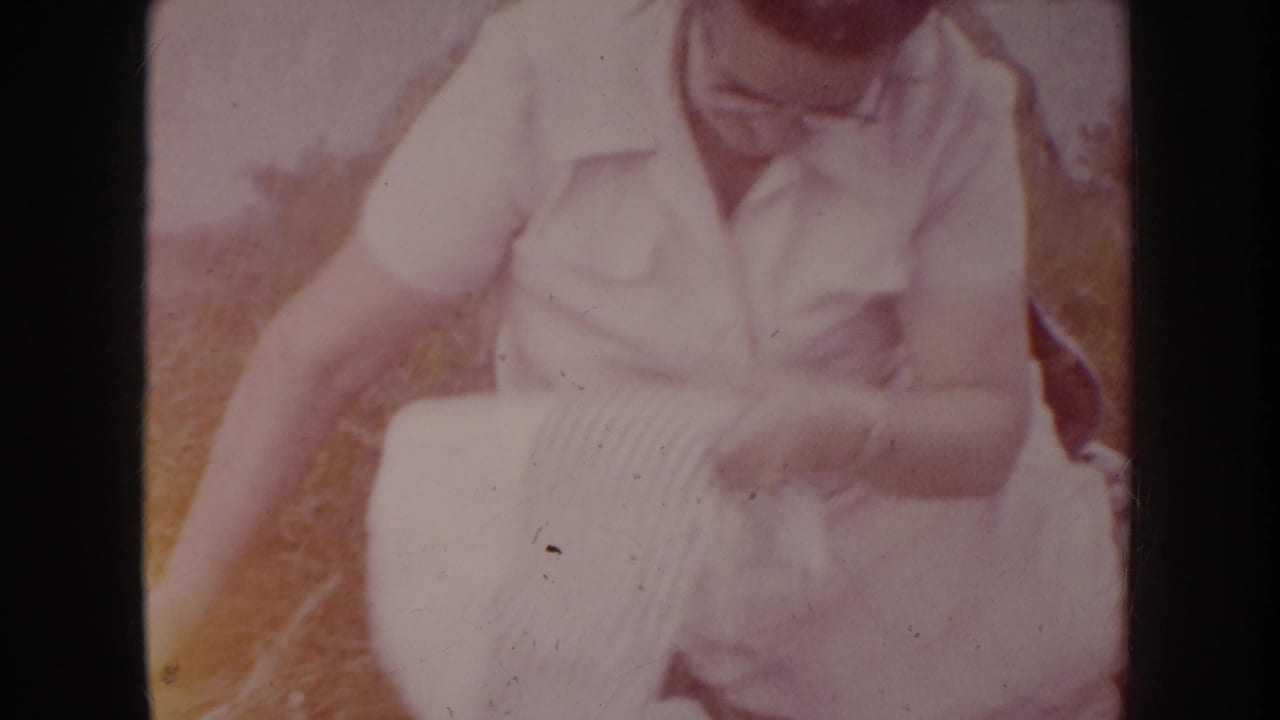 Mid-1950s woman in light short-sleeve blouse and glasses seated outdoors on dry grass, adjusting a horizontally striped cloth, South Dakota, circa 1955.
