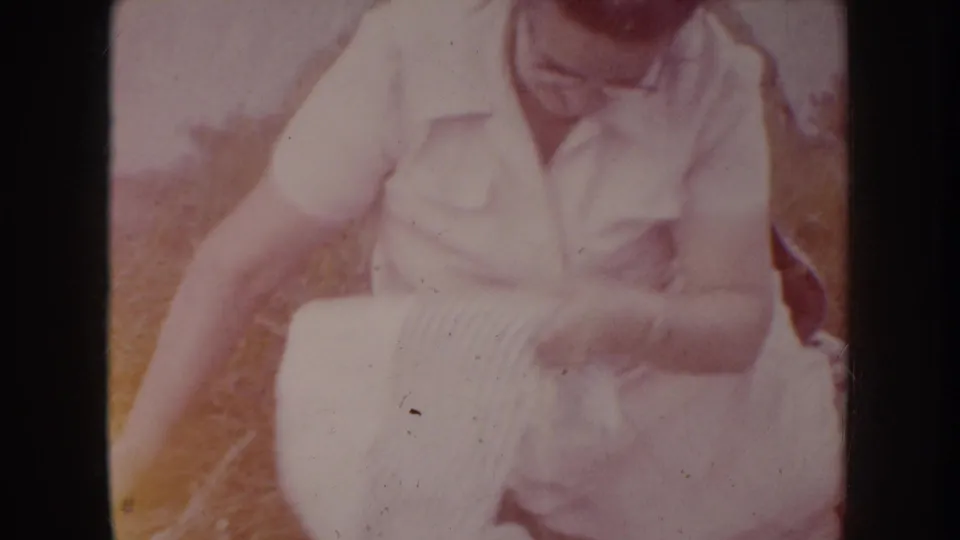 Mid-1950s woman in light short-sleeve blouse and glasses seated outdoors on dry grass, adjusting a horizontally striped cloth, South Dakota, circa 1955.