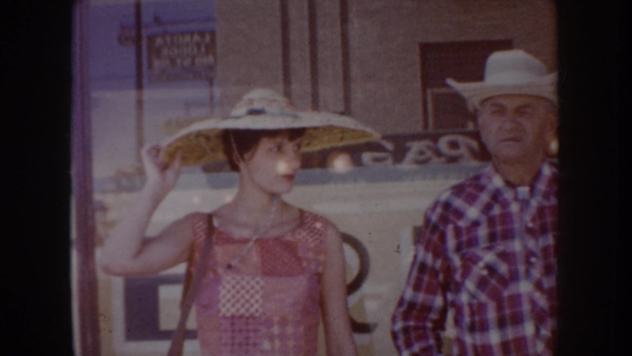 Young woman in wide-brimmed straw hat adjusts her hat; older man in white cowboy hat and plaid shirt stands beside her in front of a storefront.