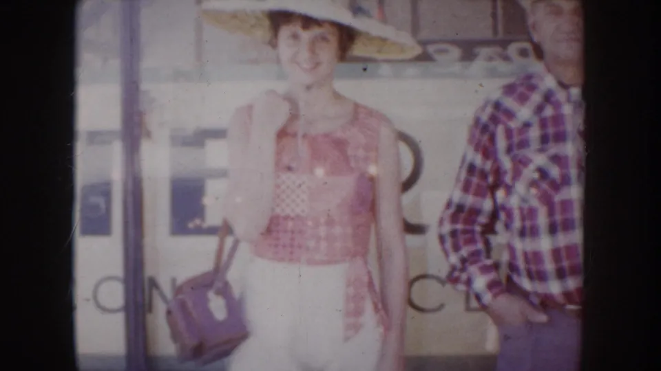 1950s color 8mm: young woman in straw hat and patchwork top smiles toward camera; older man in plaid shirt stands right; partial storefront lettering visible, South Dakota, c. 1955.