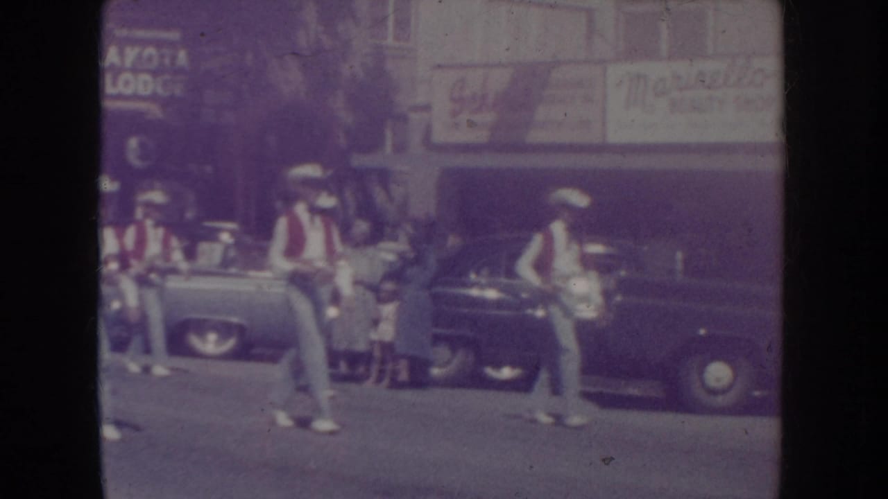 8mm home movie frame of a marching band in red-and-white western uniforms on a South Dakota parade street, circa 1955, with period automobiles and partial storefront signage