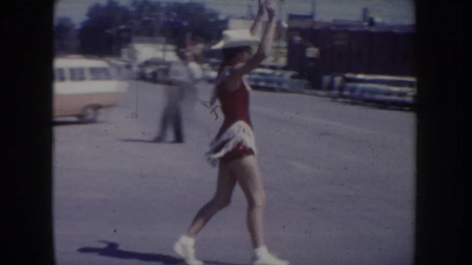 8mm home movie frame of a female dancer in red top, white fringed skirt, and cowboy hat mid-performance in a South Dakota street parade, circa 1955, with period automobiles behind her