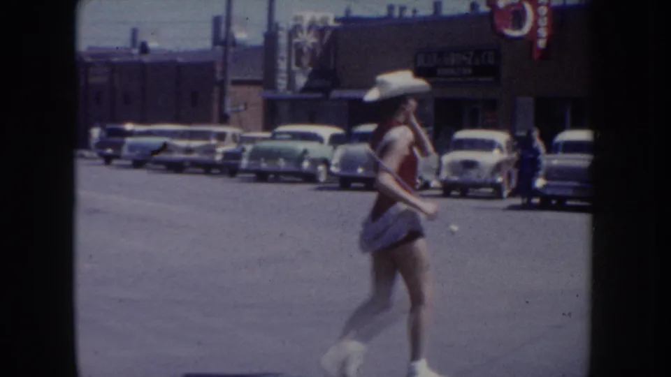 8mm home movie frame of a parade dancer in western costume passing parked period automobiles in South Dakota, circa 1955, with a brick building and red-white-blue bunting visible in the background