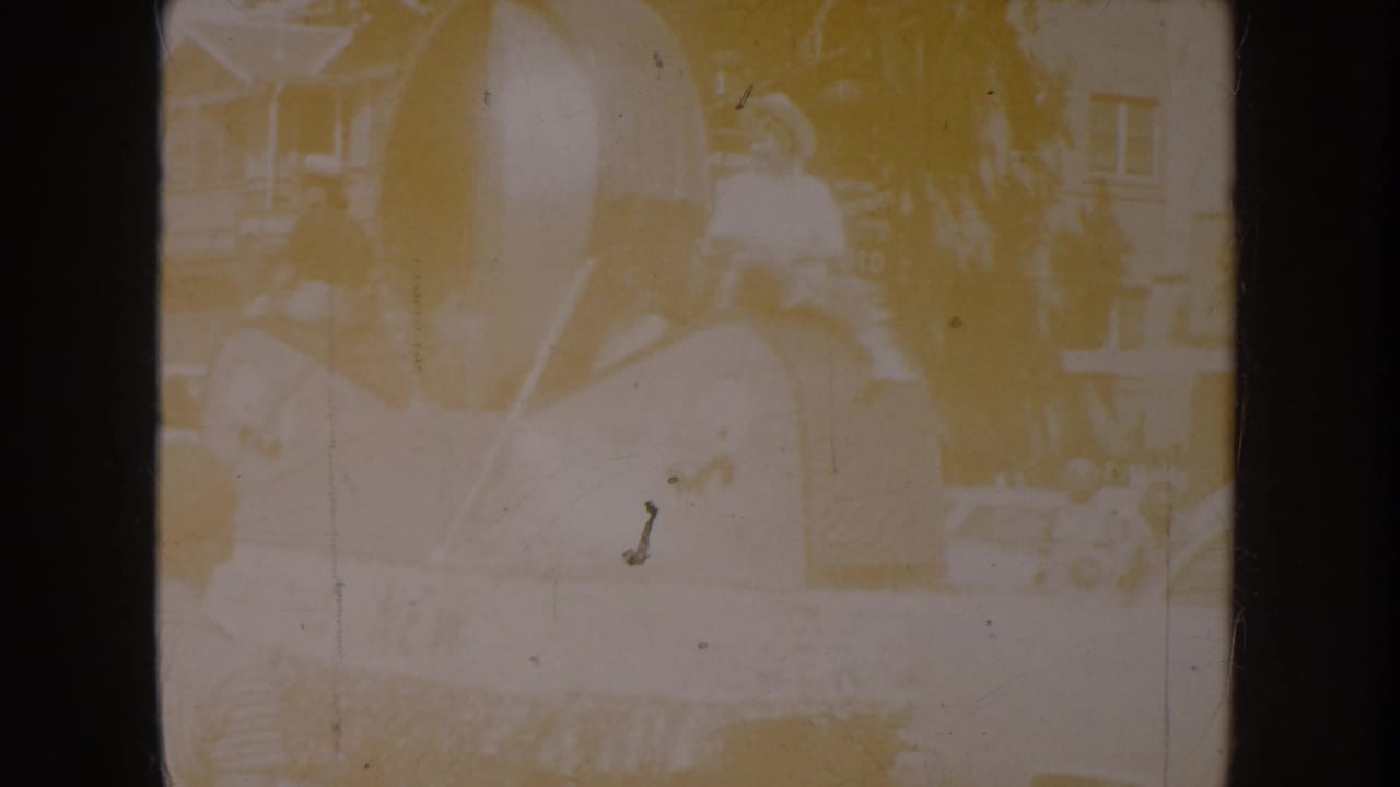 Heavily yellowed and overexposed 8mm frame showing faint figures near what may be a vehicle, with visible scratches and film grain.