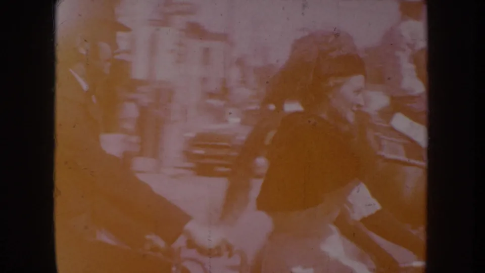 Overexposed mid-clip frame from a circa-1955 8mm parade recording; two figures visible through heavy orange dye-shift, possibly cyclists or parade participants.