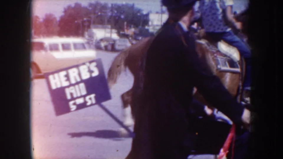 Horseback rider in dark suit and hat passes a 'HERB'S / 5TH ST' storefront sign during a circa-1955 street parade in South Dakota; 1950s vehicles line the street behind.
