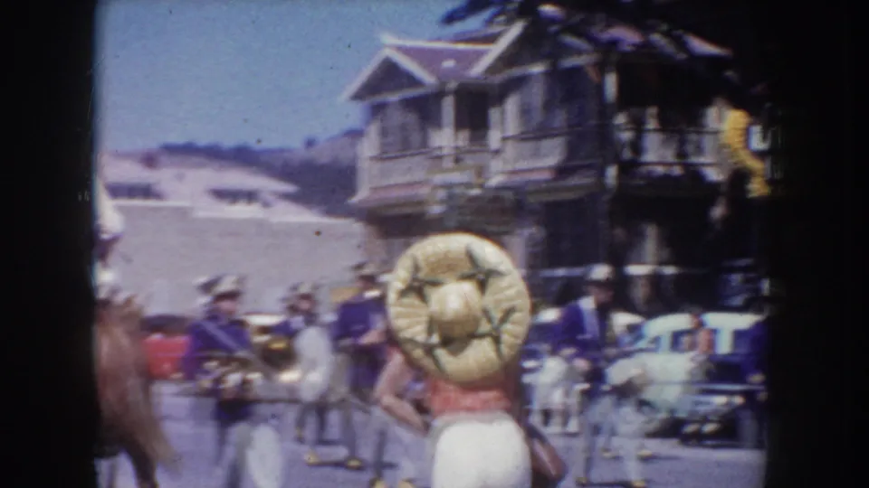 Crowd spectator wearing an oversized straw hat viewed from behind during a circa-1955 street parade; a two-story clapboard building and clear blue sky are visible in the background.