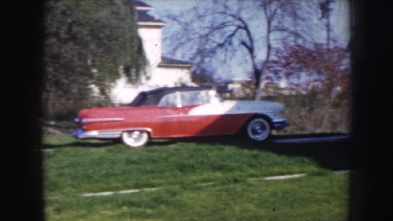 Red and white two-tone convertible parked on a green grass lawn beside a white house, bare deciduous trees in the background.