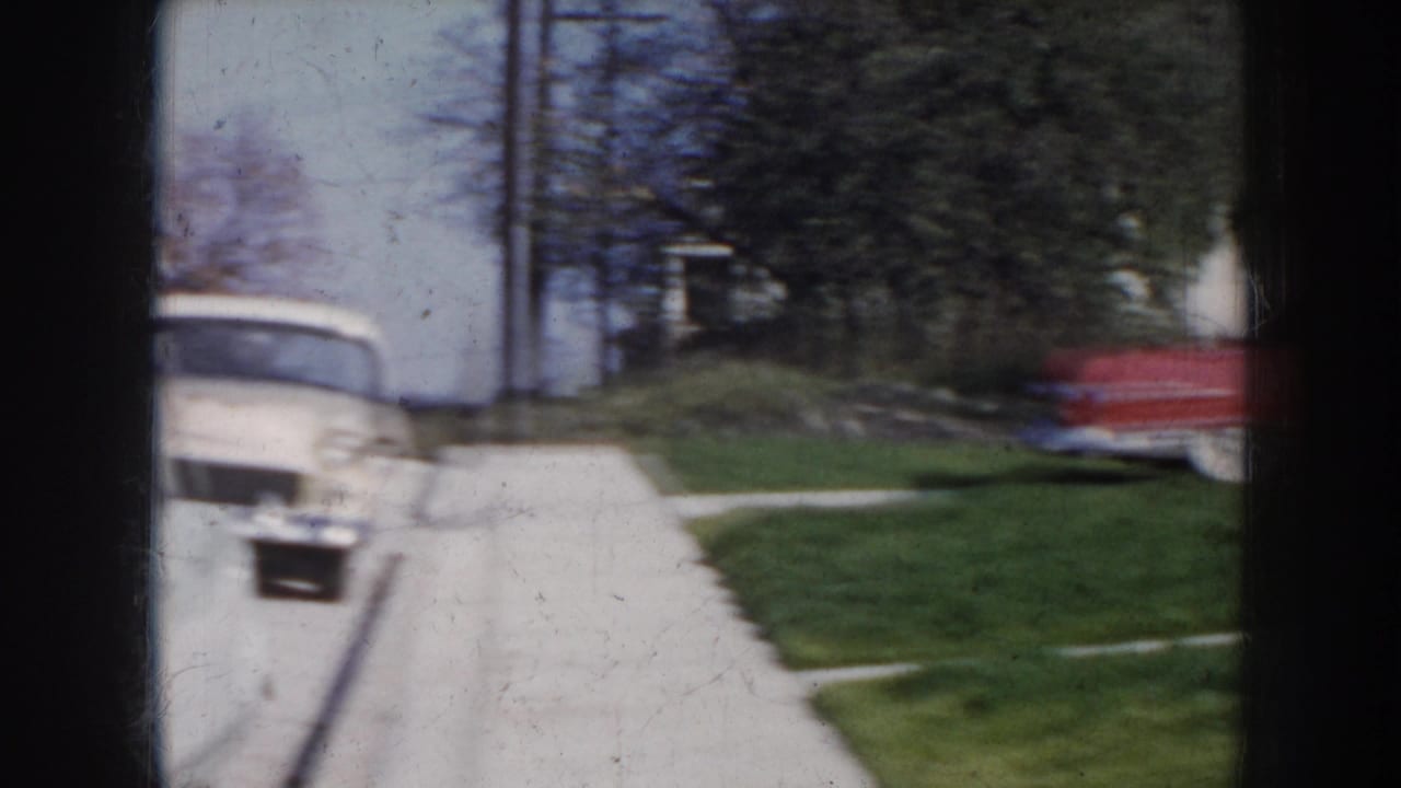 Wide pan across a concrete driveway with a light-colored vehicle in center and the red convertible visible at the right frame edge.