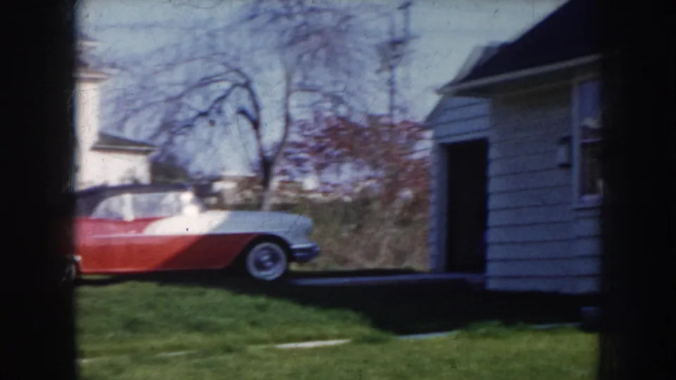 Camera panning from the convertible toward the white clapboard house exterior with bare-branched trees, Nebraska suburban scene circa 1956.