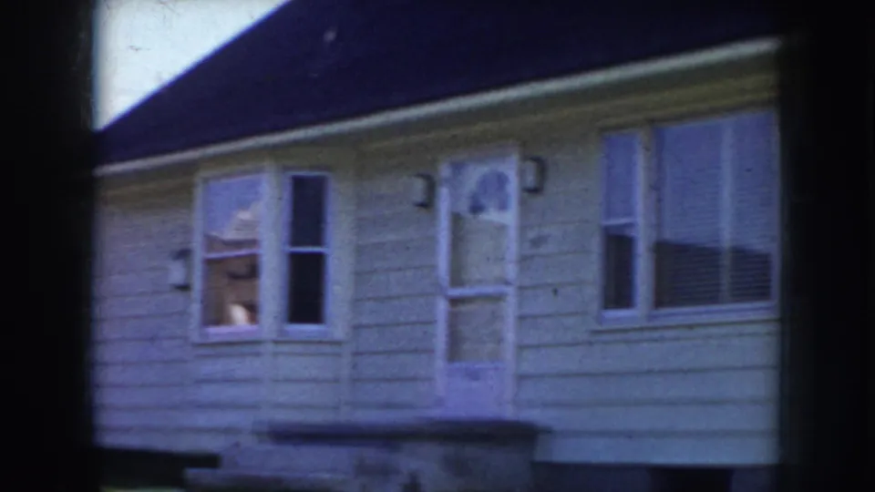 Close framing of white clapboard house windows and exterior wall; a possible figure appears through one window, Nebraska suburban home circa 1956.