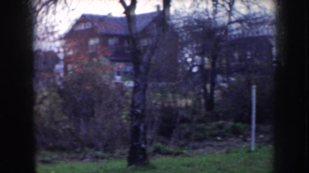 8mm home movie frame showing bare trees, a grassy embankment, and substantial two-story residential houses in a quiet neighborhood, consistent with mid-1950s Nebraska.