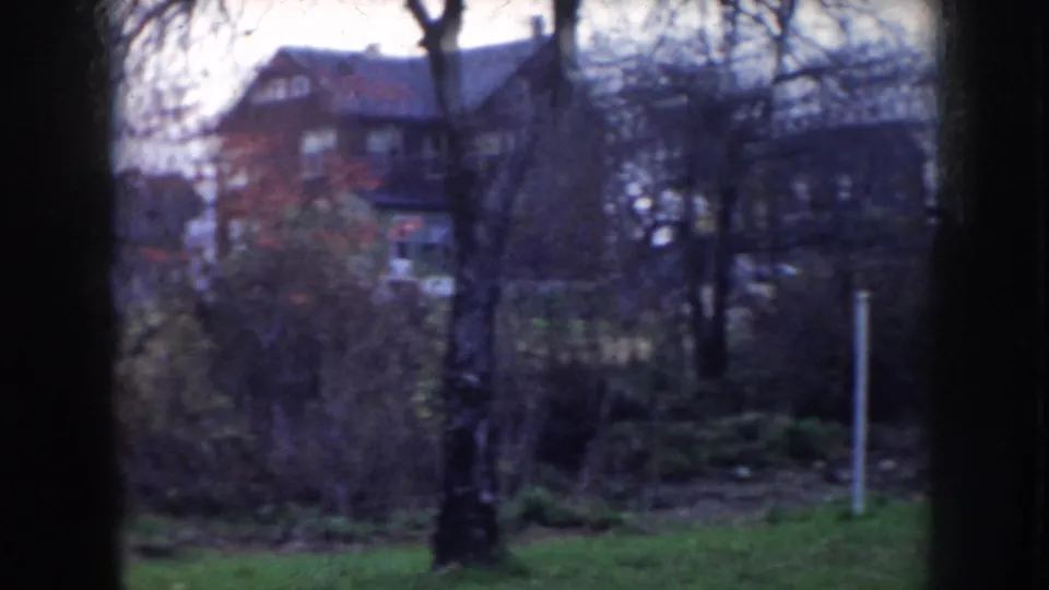 8mm home movie frame showing bare trees, a grassy embankment, and substantial two-story residential houses in a quiet neighborhood, consistent with mid-1950s Nebraska.