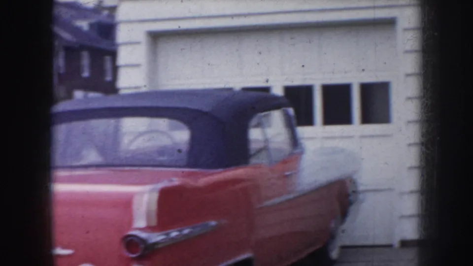 Red 1950s American convertible — tail fins and raised black soft top clearly visible — parked in a residential driveway beside a white-sided garage, consistent with a mid-1950s Nebraska suburban setting.