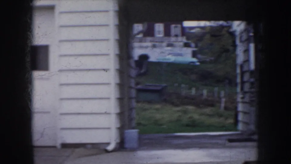 View past a white-sided residential structure to the neighborhood beyond, with bare trees, green lawn, and what appears to be a parked vehicle partially visible through the gap, circa 1956 Nebraska.
