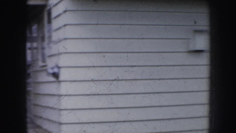 1956 Nebraska 8mm frame with a close, slightly blurred view of pale clapboard siding, window edges, exterior fixtures, and a dark vertical porch post dividing the composition.