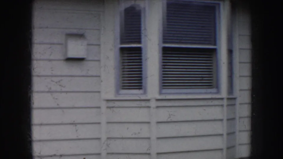 1956 Nebraska 8mm frame showing a pale house facade with two windows, visible blinds, horizontal siding, trim, and soft grain from the home-movie scan.