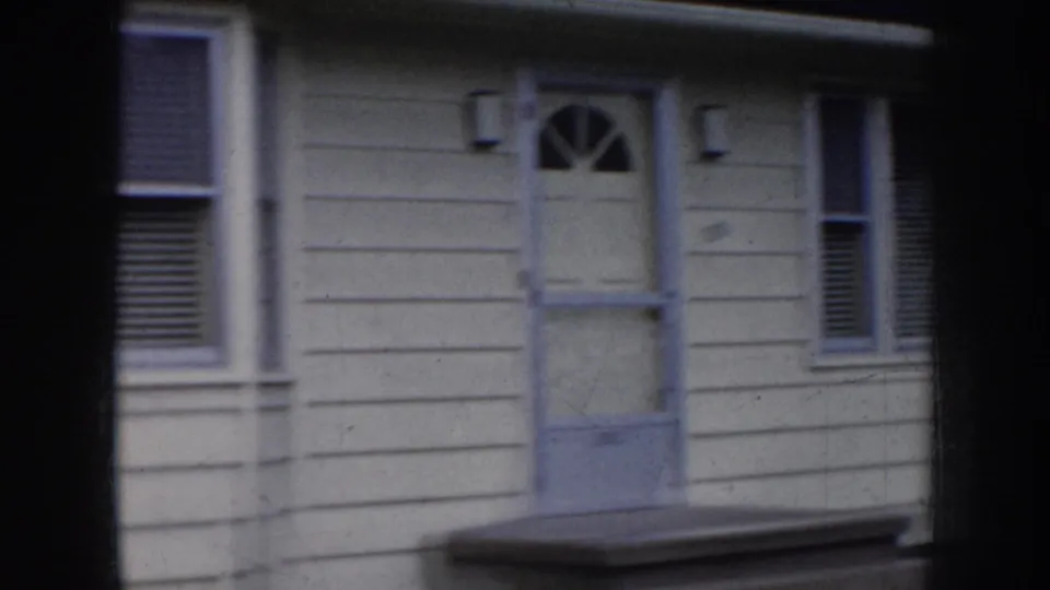1956 Nebraska 8mm frame of a pale house entrance with a worn door, fanlight-style upper window, porch step, adjacent windows, siding, and dark vignetting at the scan edges.