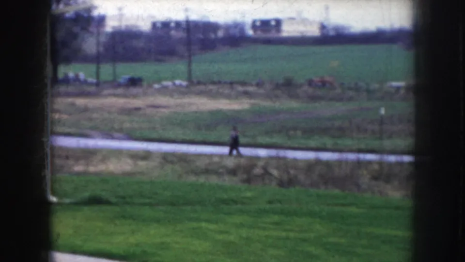8mm frame showing rural-suburban edge, Nebraska c.1956 — green field, paved path, lone pedestrian, and utility poles stretching toward the horizon.