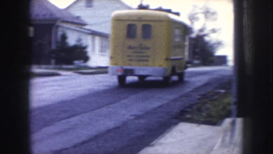 Yellow commercial van on a suburban Nebraska street, c.1956 — vehicle has visible text or livery on its rear panel; white house and concrete sidewalk visible.