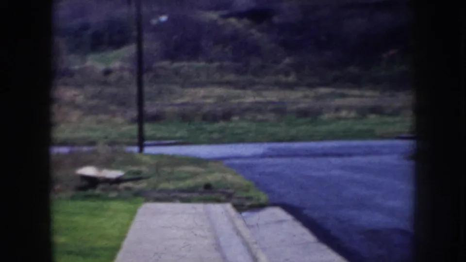 8mm frame from Nebraska, c.1956 — paved sidewalk edge with grass strip, utility pole, and open transitional lot or field; no people visible.