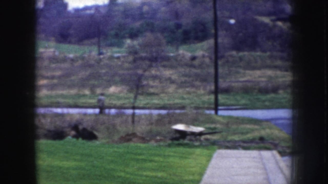 8mm home movie view through a window in Nebraska, circa 1956, showing a rural yard, paved road, and a pedestrian at road distance.