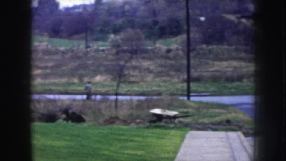 8mm home movie view through a window in Nebraska, circa 1956, showing a rural yard, paved road, and a pedestrian at road distance.