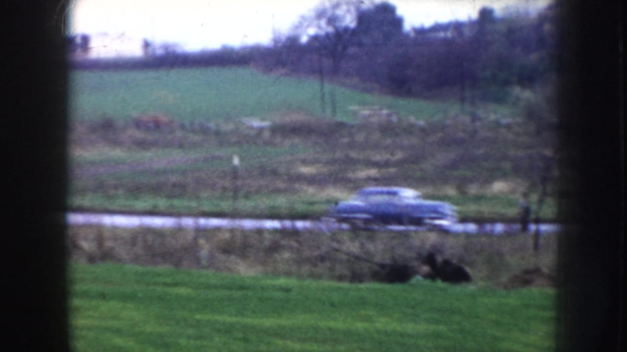 View through window showing a dark 1950s sedan passing along a rural road with green fields and trees beyond.