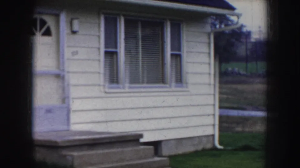 Mid-century American house facade in Nebraska, circa 1956, showing cream horizontal siding, a door with fan-window surround, concrete porch steps, and an exterior light fixture.