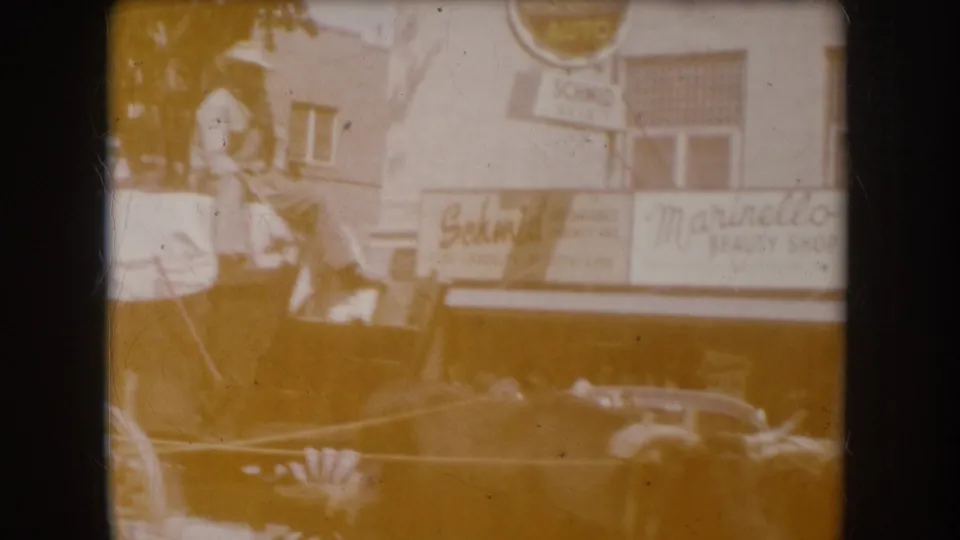 8mm film showing heavy amber dye-fade degradation, circa 1956 South Dakota — a figure stands near the wagon while 'Schmid' and 'Marinello' signs remain faintly visible through the color cast.
