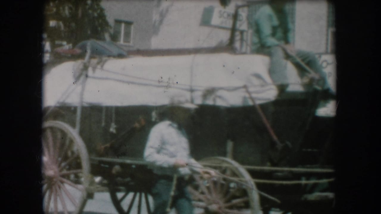 Opening 8mm home movie frame showing a horse-drawn buggy with a covered or canopied top on a small-town street, likely South Dakota circa 1956; a figure in white visible in the carriage seat.