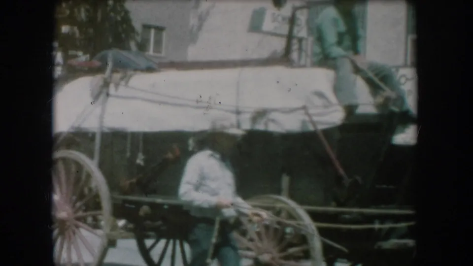 Opening 8mm home movie frame showing a horse-drawn buggy with a covered or canopied top on a small-town street, likely South Dakota circa 1956; a figure in white visible in the carriage seat.