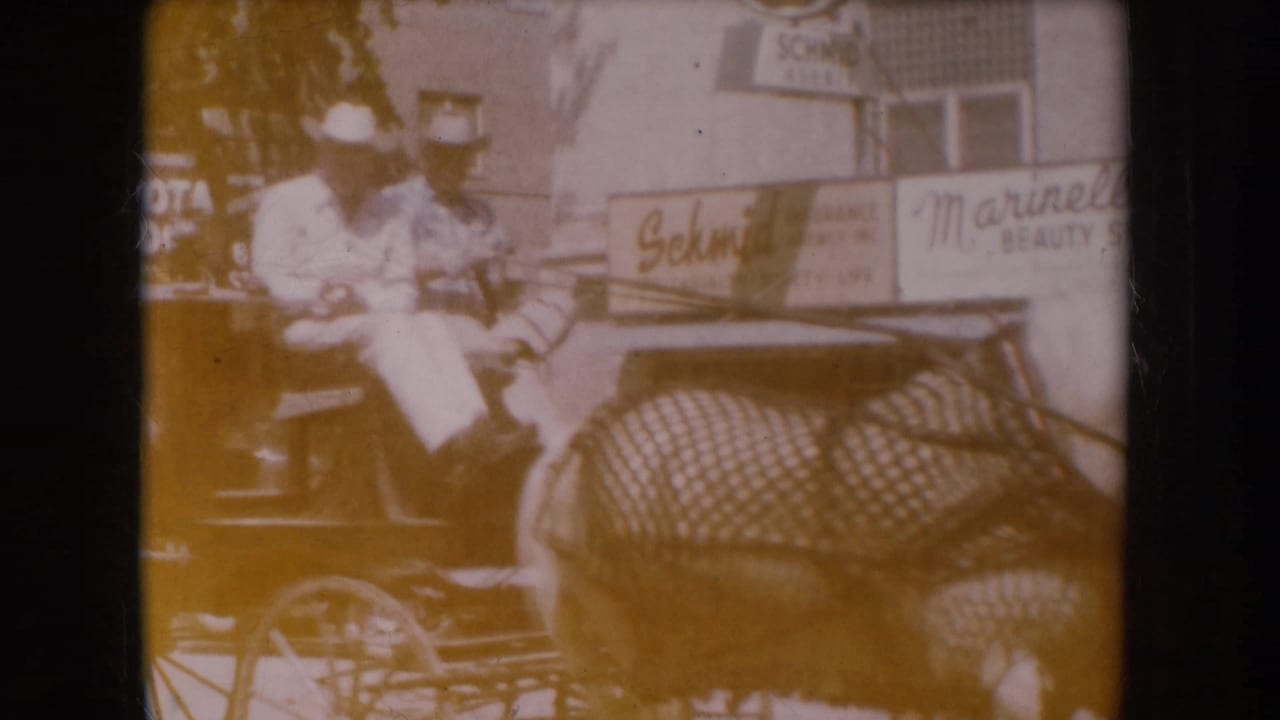 Amber-shifted 8mm frame: man in cowboy hat and white shirt seated in a buggy; partial Schmid and Marinello Beauty signs legible on storefronts behind him.