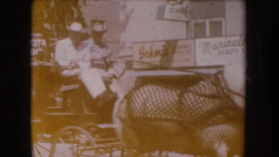 Heavily amber-degraded 8mm frame showing a man in white shirt and cowboy hat in a horse-drawn buggy; 'Schmid' and 'Marinello Beauty' storefront signs partially legible in the background, likely South Dakota circa 1956.