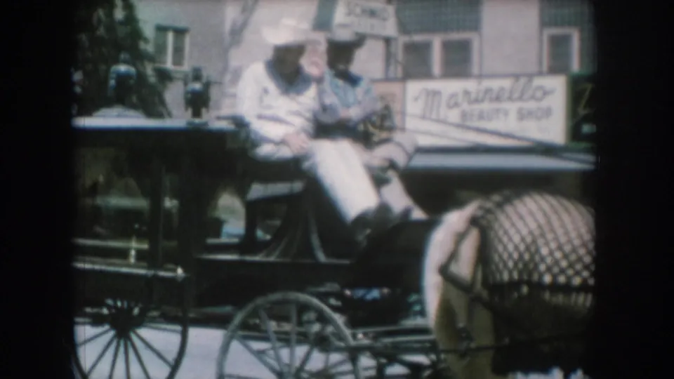 8mm home movie frame circa 1956, likely South Dakota: man in white shirt and cowboy hat rides an open buggy past a 'Marinello Beauty Shop' storefront; partial Schmid sign also visible on brick facade.