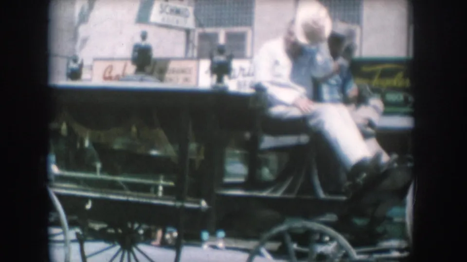 8mm frame circa 1956, likely South Dakota: two riders in an open horse-drawn buggy passing mid-century commercial storefronts; partial Schmid and Marinello signs visible; brighter exposure than preceding frames.