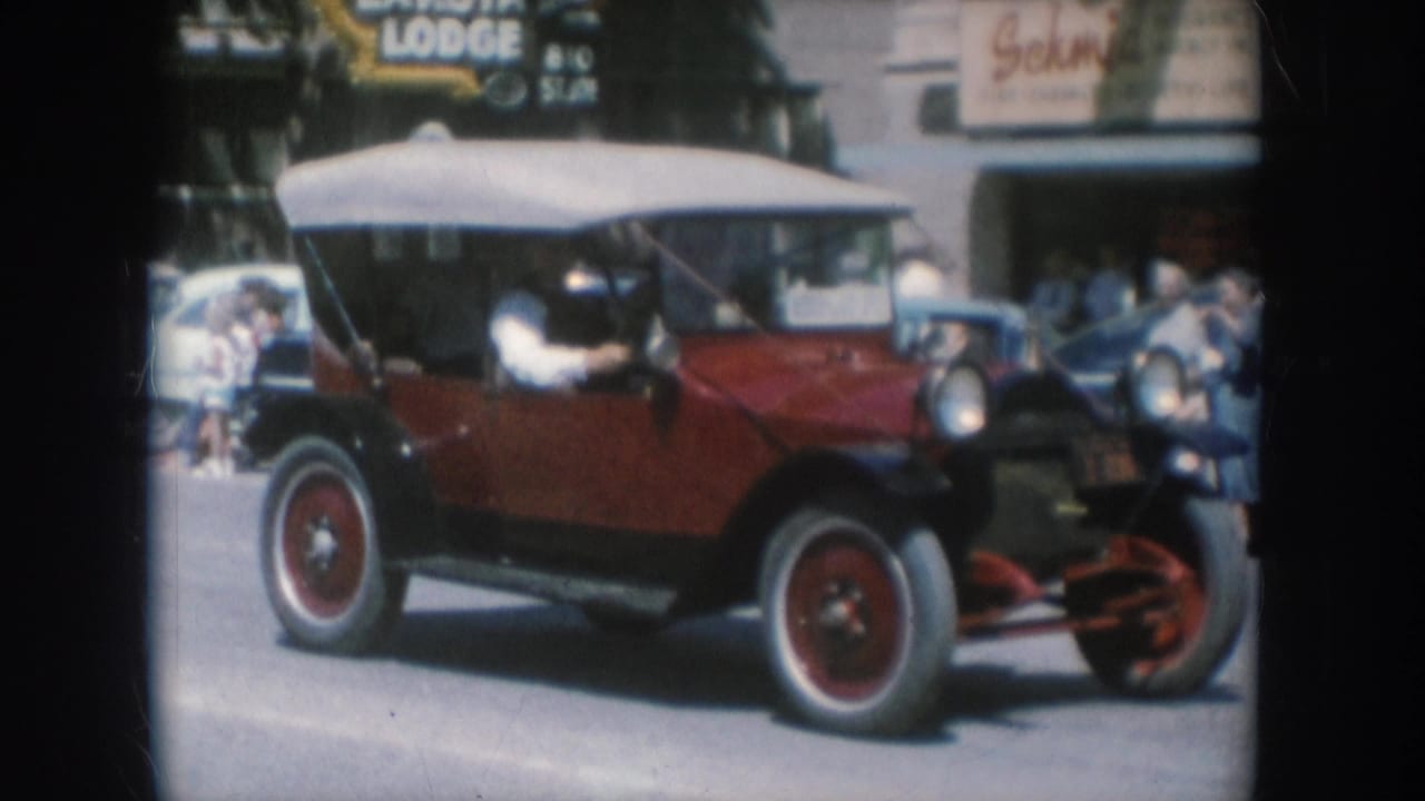 Early-1920s-era red open touring car with canvas top and spoke wheels on a South Dakota small-town street, 1956; LAKOTA LODGE sign partially visible at upper left.