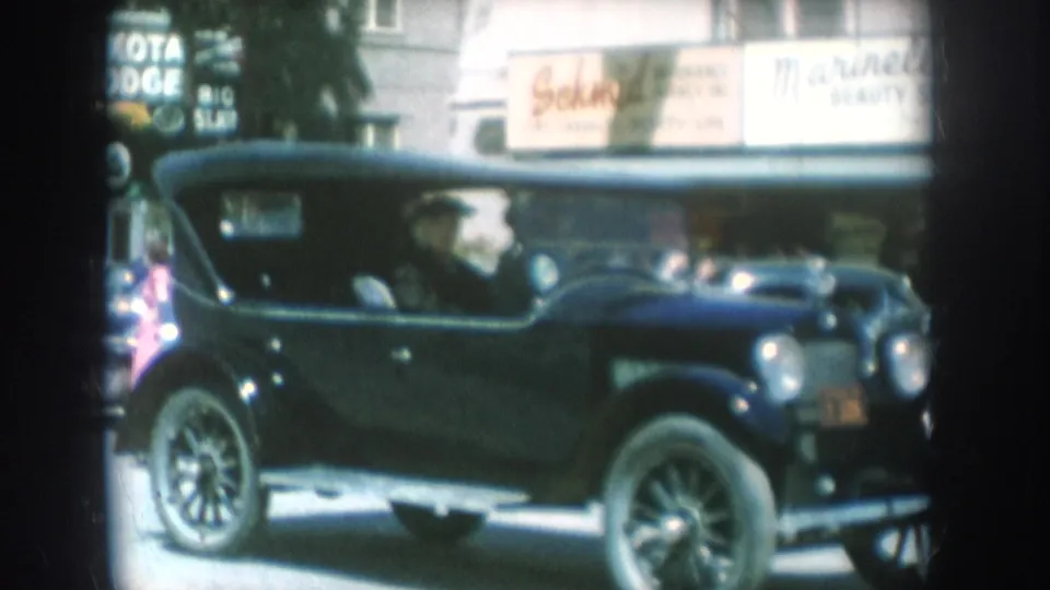 Black vintage touring car passes South Dakota storefronts including a partial Lakota Lodge sign and a beauty salon sign, with a spectator visible at the left edge, circa 1956.