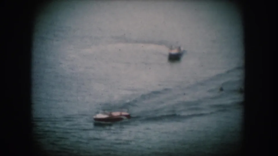 Two small open motorboats cruise a lake in what appears to be 1950s home movie footage from South Dakota; the lower vessel shows warm coloring consistent with recreational boat finishes of the era.