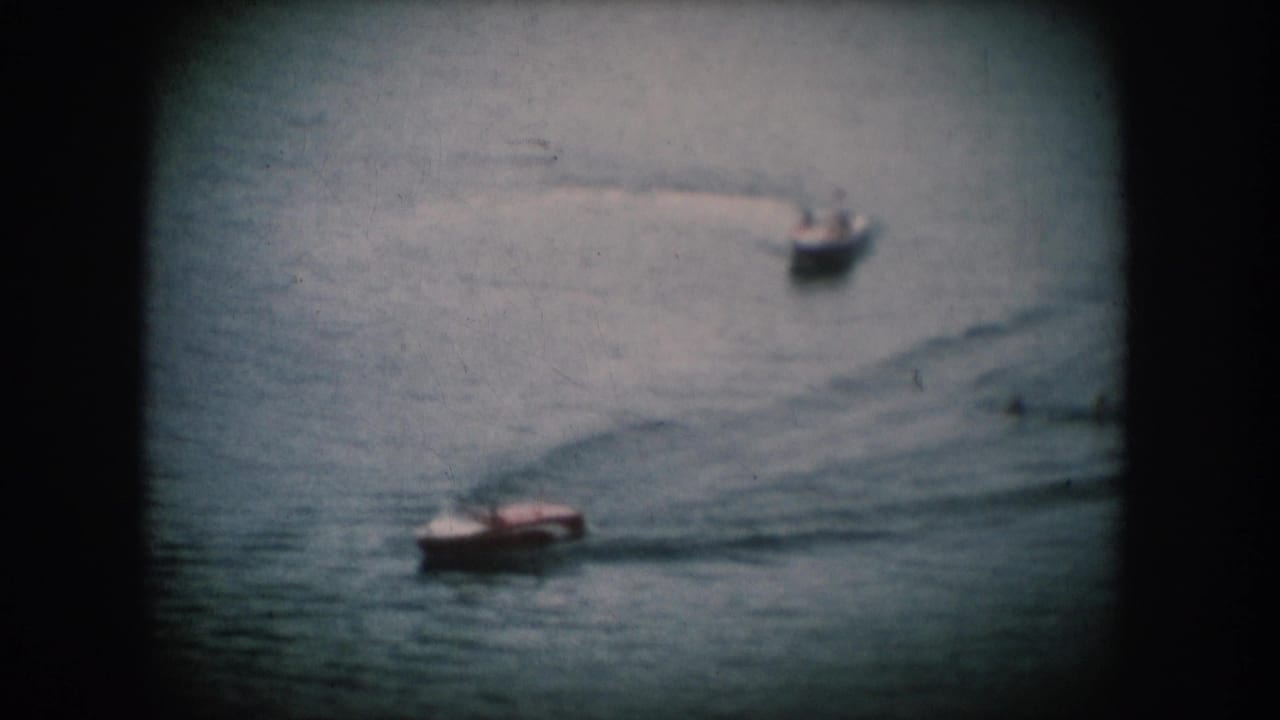 Two small motorboats cross a calm lake surface in what may be South Dakota, c. 1956; blue-tinted 8mm footage with rounded projector vignette.