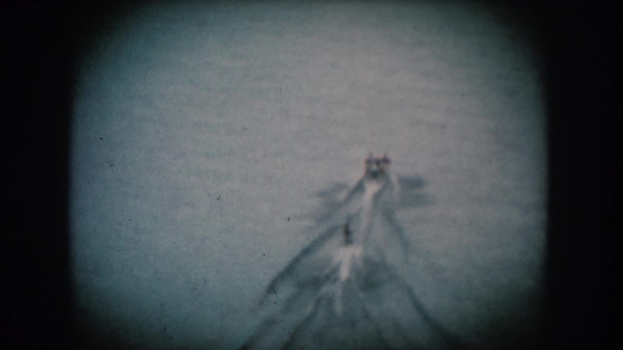 Single motorboat centered on open water leaving a V-shaped wake; blue-tinted 8mm film with circular vignette.